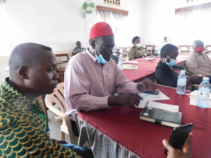 Due to the Covid-19 pandemic, Emily Sample conducts her research over Zoom. Two Ugandan men are shown sitting at a table and talking to Sample via Zoom on an iPhone. 