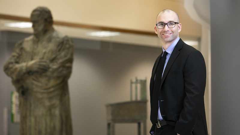 Adam Crepelle stands in the law school's atrium, wearing a black suit and light blue collared shirt with his hands in his pockets. He is wearing dark, thick-rimmed glasses and smiling at the camera.