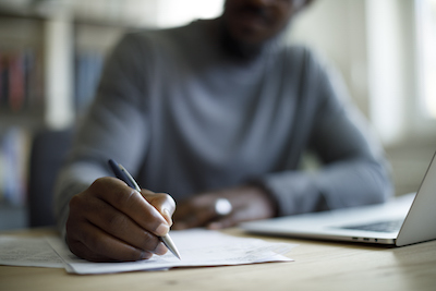man writing with pen by a laptop