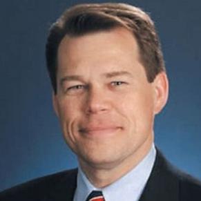 A man in a dark suit with a blue shirt and a striped tie smiles at the camera.