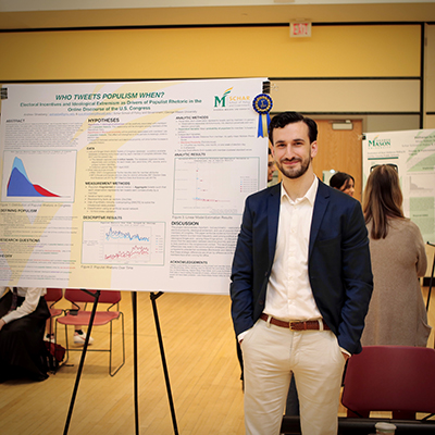 A man with a dark beard and jacket stands in front of a poster on a stand.