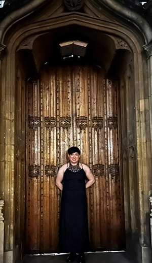 A woman with short dark hair smiles broadly as she stands in the doorway of an ancient stone building.