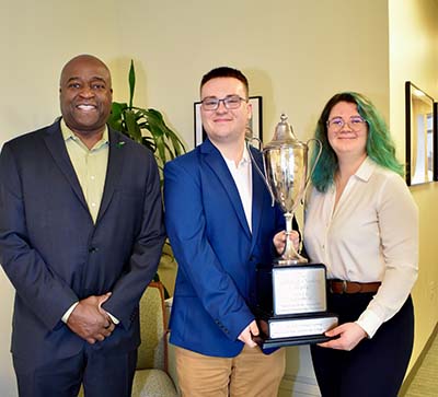 A man in a blue jacket with his hands folded in front of him stands next to a man in a blue jacket and a woman with green hair holding a large trophy.