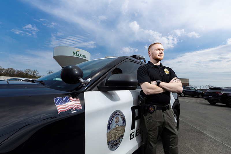 A man in a police uniform and stands next to a police car with the Mason water tower behind it.