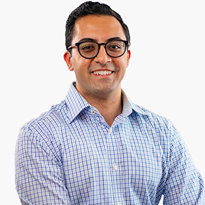 A man with dark hair and black eyeglasses wears a blue checked shirt and smiles at the camera.