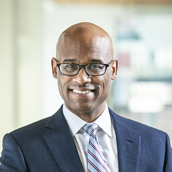 Smiling man wearing black glasses and dark suit with striped tie.