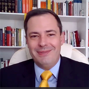 A man with short dark hair and wearing a dark jacket, blue shirt, and gold necktie is captures in a Zoom screenshot.
