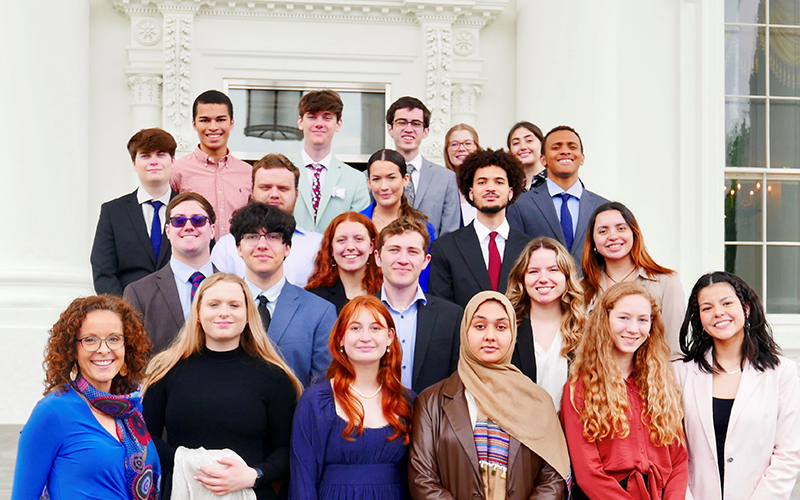 A group of smiling young adults and a woman in a blue dress stand on steps in front of a white house.