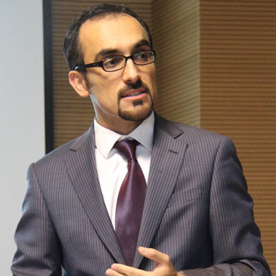 A man in a gray suit with stripes and a purple tie and eyeglasses looks to his left.