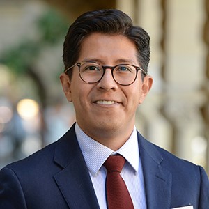 A man wearing eyeglasses and a blue suit with red tie smiles at the camera.