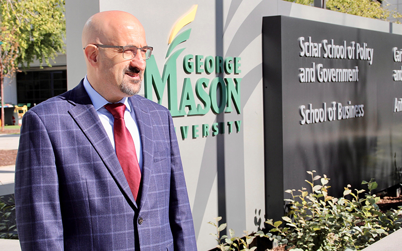 A bald man in eye glasses and a blue suit stands in front of a George Mason University sign.