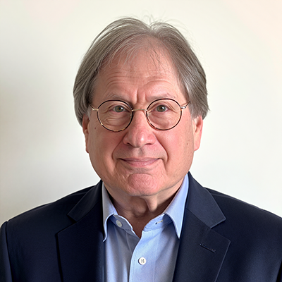 A man with glasses, a black suit, and blue collared shirt stands and smiles