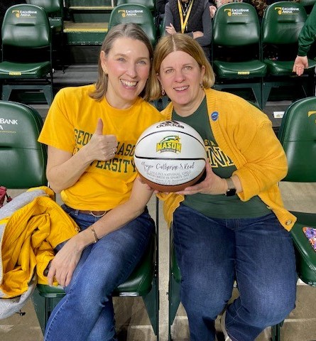 Traci Kendall and Fairfax City Mayor Catherine Read sit together at a George Mason basketball game. Read is holding a personalized basketball