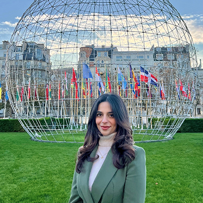 A young woman with dark hair and wearing a green jacket stands in front of a skeletal globe and flags.