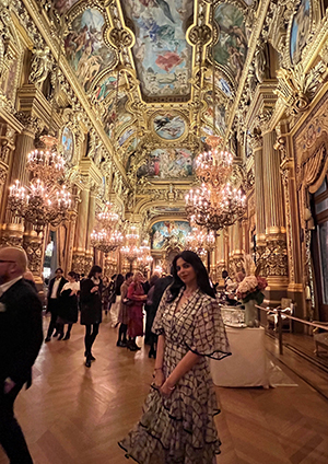 A young woman in a dress stands in an elaborate foyer with chandeliers and frescos on the ceiling.