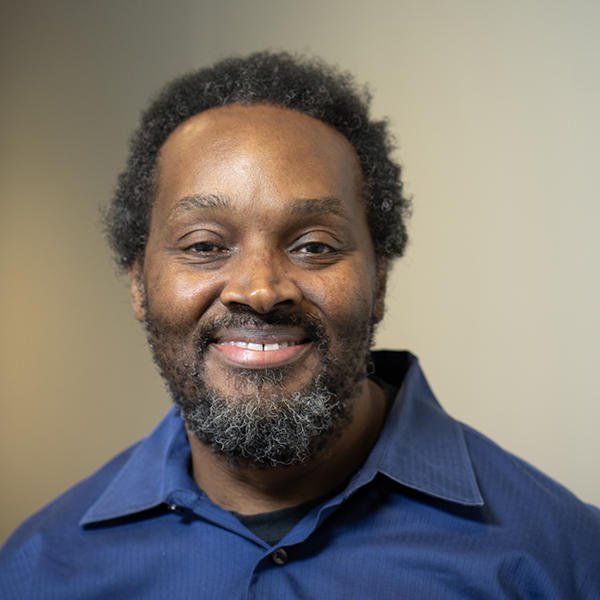 A headshot of a Black man with a beard, wearing a button down shirt, looking at the camera.